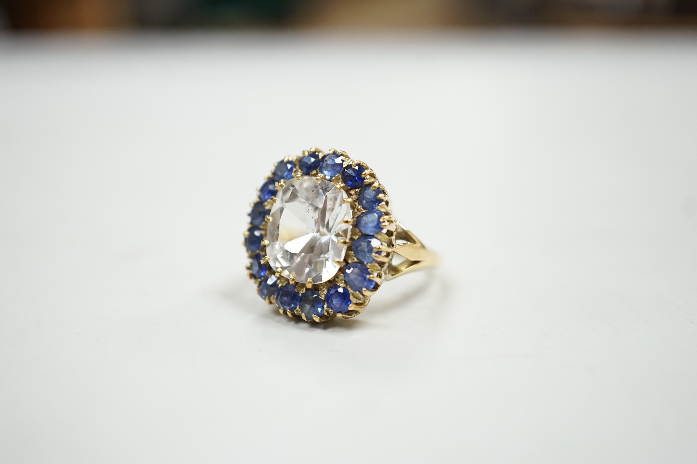 An early 20th century yellow metal and single stone cushion cut white sapphire set dress ring, bordered by round and oval cut blue sapphires, size P, gross weight 9.4 grams.